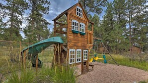A wooden playhouse with a slide and swing set, surrounded by trees and a gravel area. - Stunning Evergreen Chalet w/ Private Hot Tub! (Evergreen)