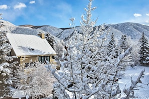 Uma casa com telhado inclinado, rodeada por árvores cobertas de neve e montanhas ao fundo.