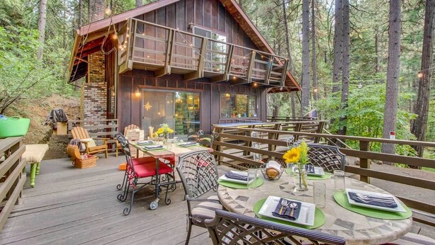 A wooden cabin with a deck, furnished with tables and chairs, surrounded by a forest.