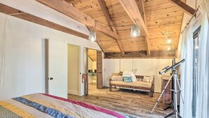 A bedroom with a wooden ceiling, a bed with a colorful quilt, a sofa, and a tripod with a telescope.