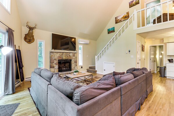 A spacious living room with a stone fireplace, a large sectional sofa, and a staircase leading to an upper level. - Furnished Deck + Fire Pit: Poconos Outdoorsy Gem (Albrightsville)