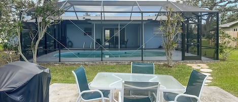 A glass-enclosed outdoor pool area with a covered patio table and chairs, surrounded by greenery.