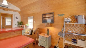 A cozy room with wooden walls, a red table, a wicker chair, a small dresser, a lamp, and a shelf with kitchen items. - Cozy Calabash Studio ~ 4 Mi to Sunset Beach! (Sunset Beach)