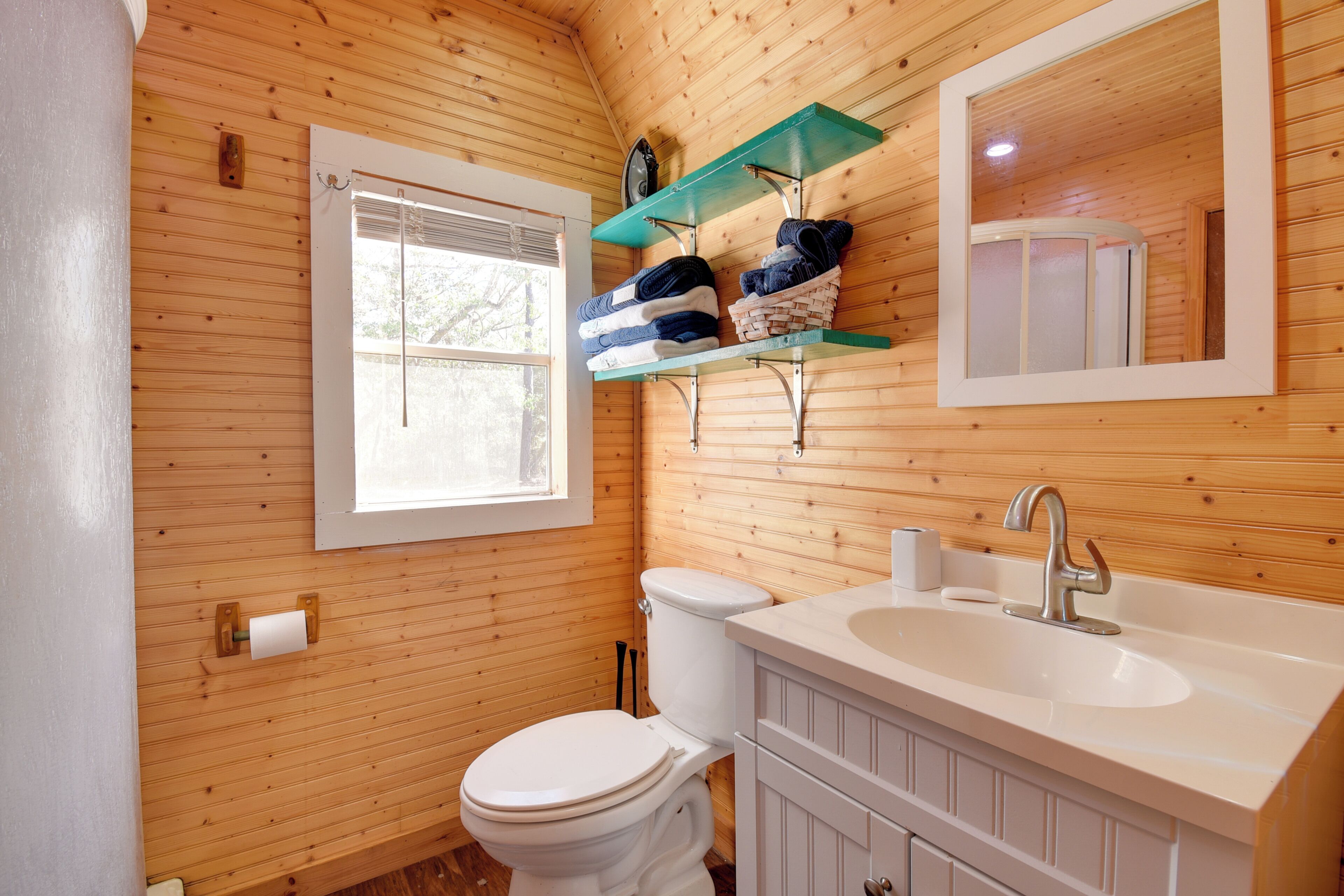 A wooden-paneled bathroom with a toilet, a sink, a mirror, and a shelf with folded towels.