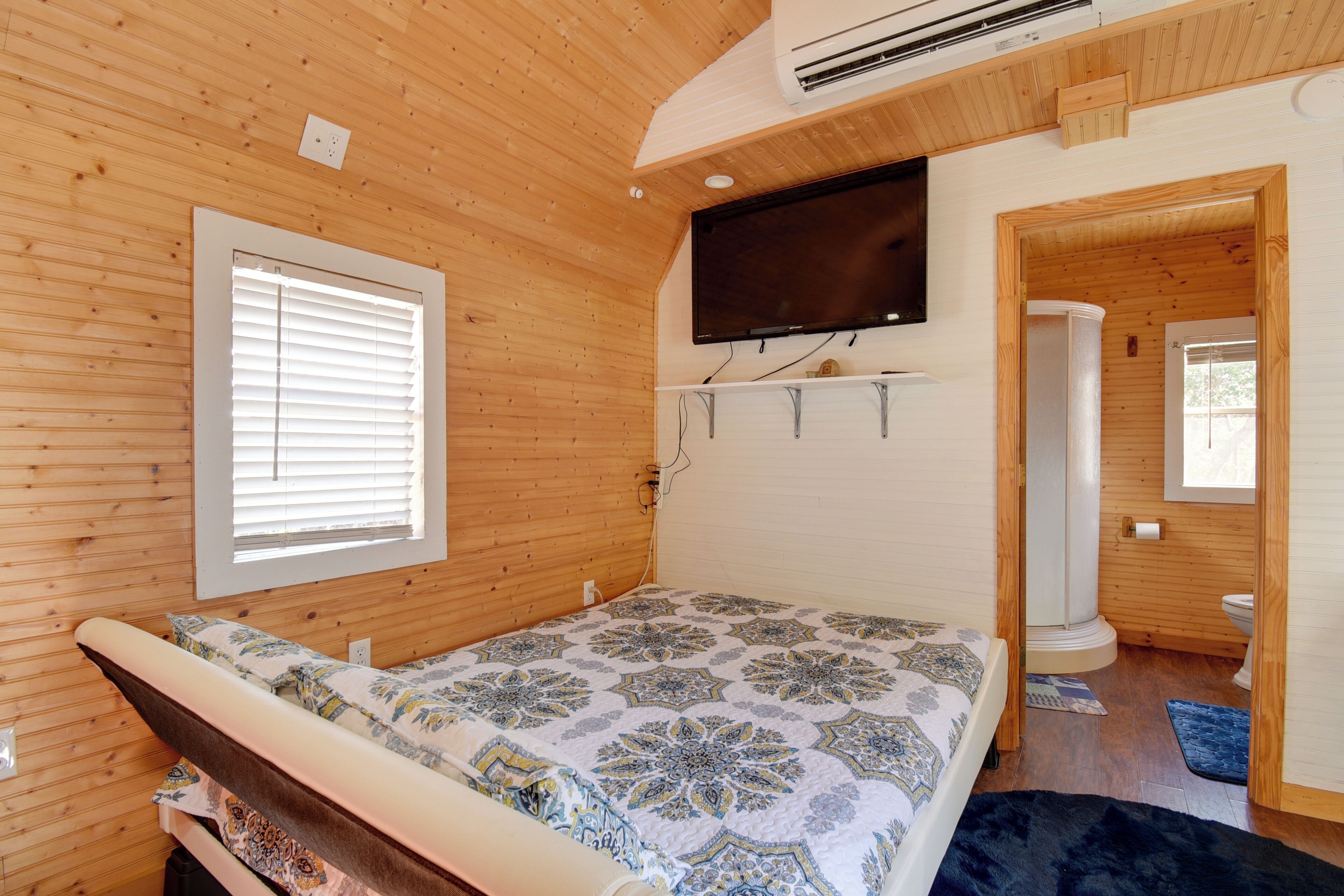 Une chambre à coucher avec un lit, un téléviseur fixé au mur, des murs en bois, une fenêtre avec des stores et une salle de bain visible à travers une porte ouverte.