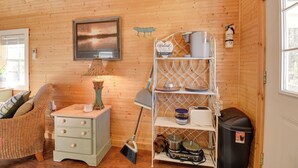 A cozy room with wooden walls, a wicker chair, a small white dresser, a lamp, a shelf with kitchen items, and a framed picture on the wall. - Cozy Calabash Studio ~ 4 Mi to Sunset Beach! (Sunset Beach)