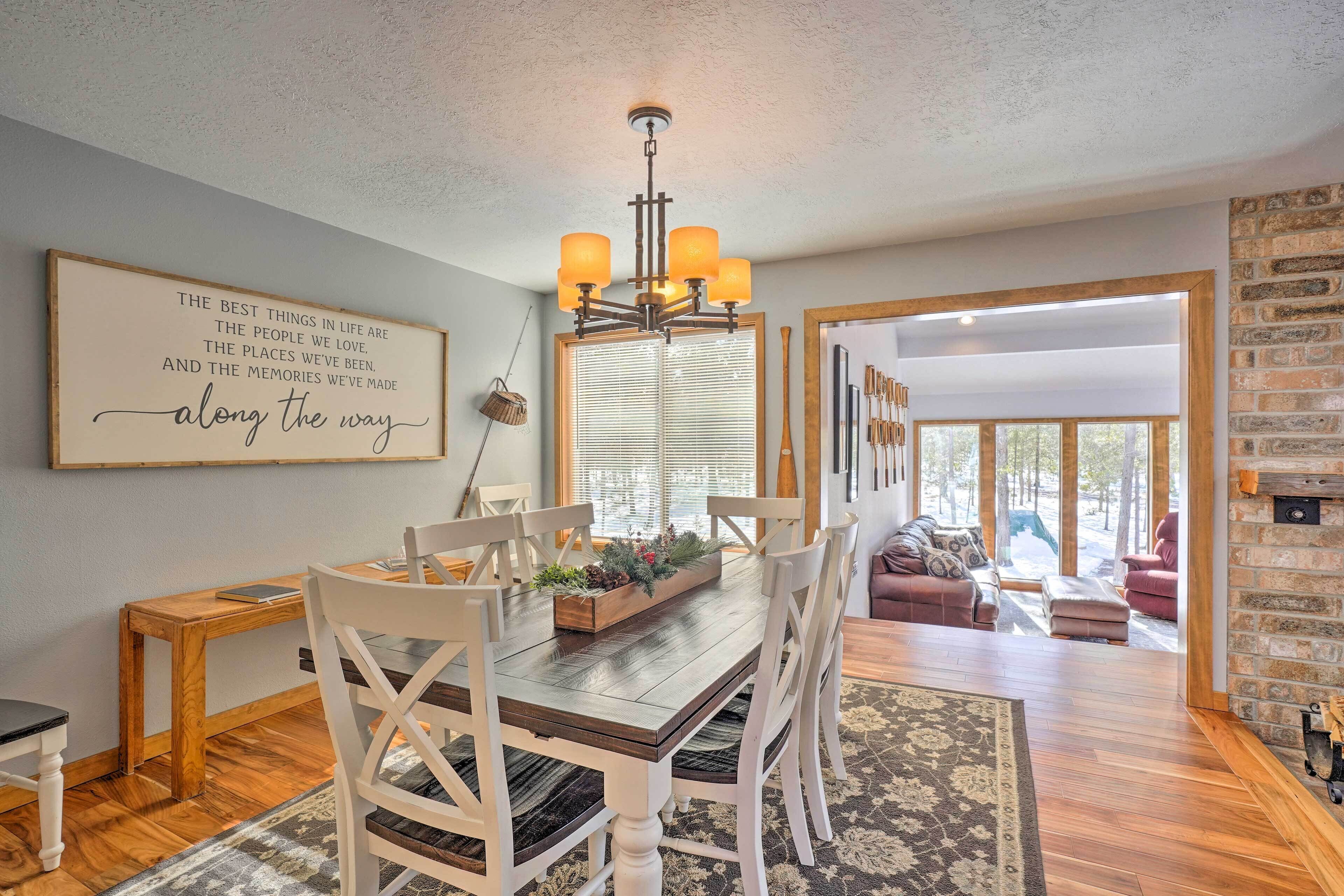 A dining area with a wooden table, white chairs, a chandelier, and a large framed quote on the wall.