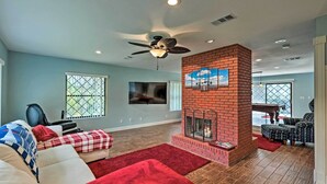 A living room with a brick fireplace, ceiling fan, and a large TV mounted on the wall. - The Aviator's Dream: Pool, Spa & Private Runway! (Port Orange)