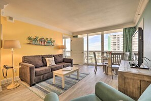 A living room with a brown sofa, a small white table, and a wooden desk with a TV. There is a balcony with a view of buildings and a door leading to an outdoor area. - Ocean-view Daytona Beach Resort Retreat w/ Balcony (Daytona Beach)