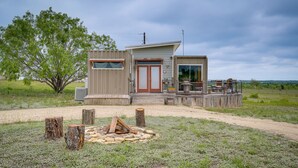 Eine kleine Holzhütte mit einer Holzterrasse, einer Feuerstelle und einem großen Fenster.