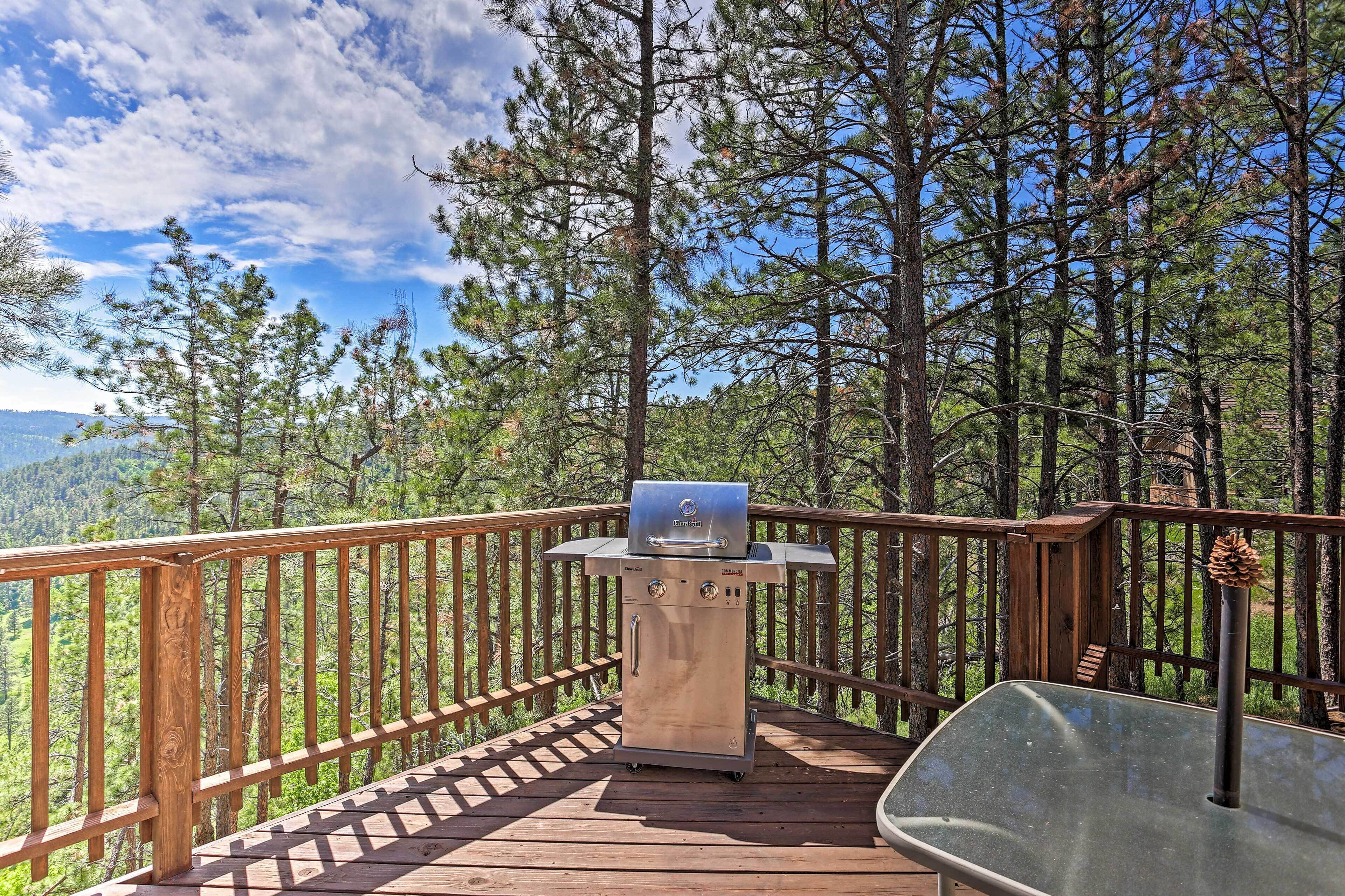 A wooden deck with a grill and a glass table, surrounded by tall pine trees and a clear sky.