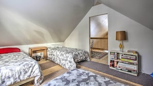 Quarto sótão pequeno e triangular com duas camas, uma estante e um abajur.