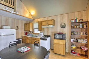 A compact kitchen with wooden cabinets, a stove, a microwave, and a dining table with a book. - Secluded Irons Cabin w/ 5-acre Yard, Deck, Grill! (Irons)