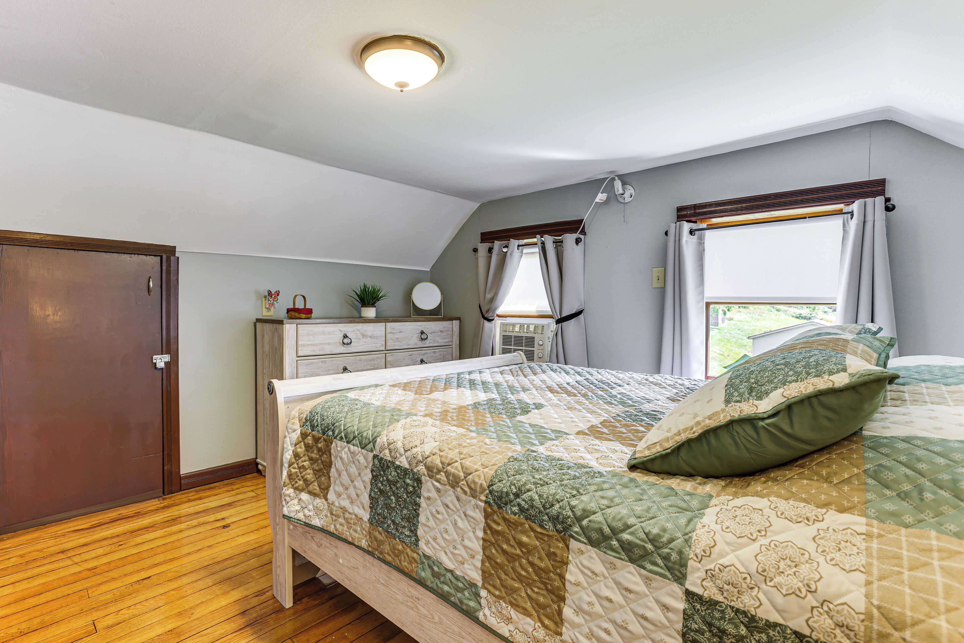 A bedroom with a wooden floor, a bed with a quilt, a dresser, a nightstand, a window with curtains, and a ceiling light.