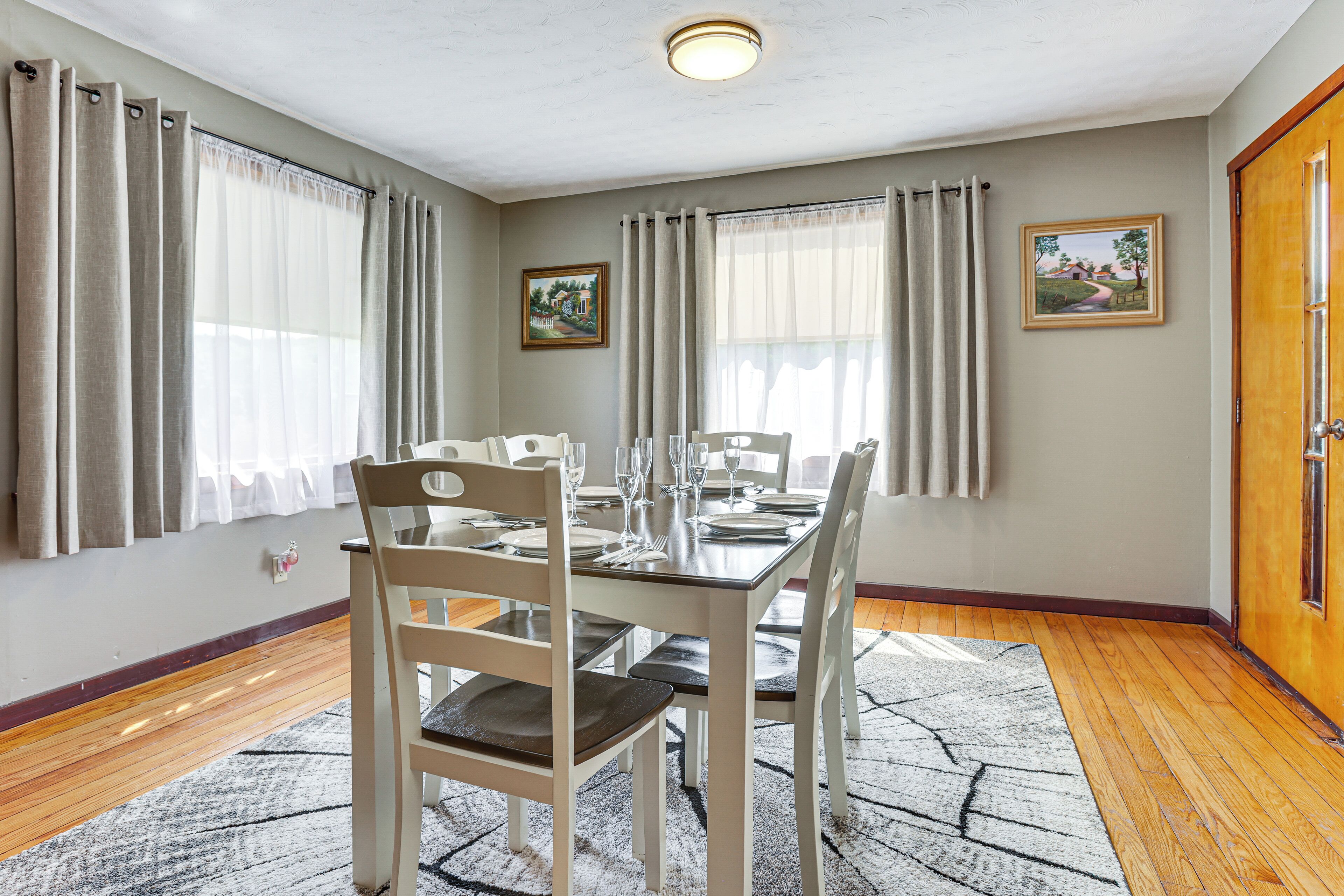 A dining room with a table set for six, a radiator, and two windows with curtains.