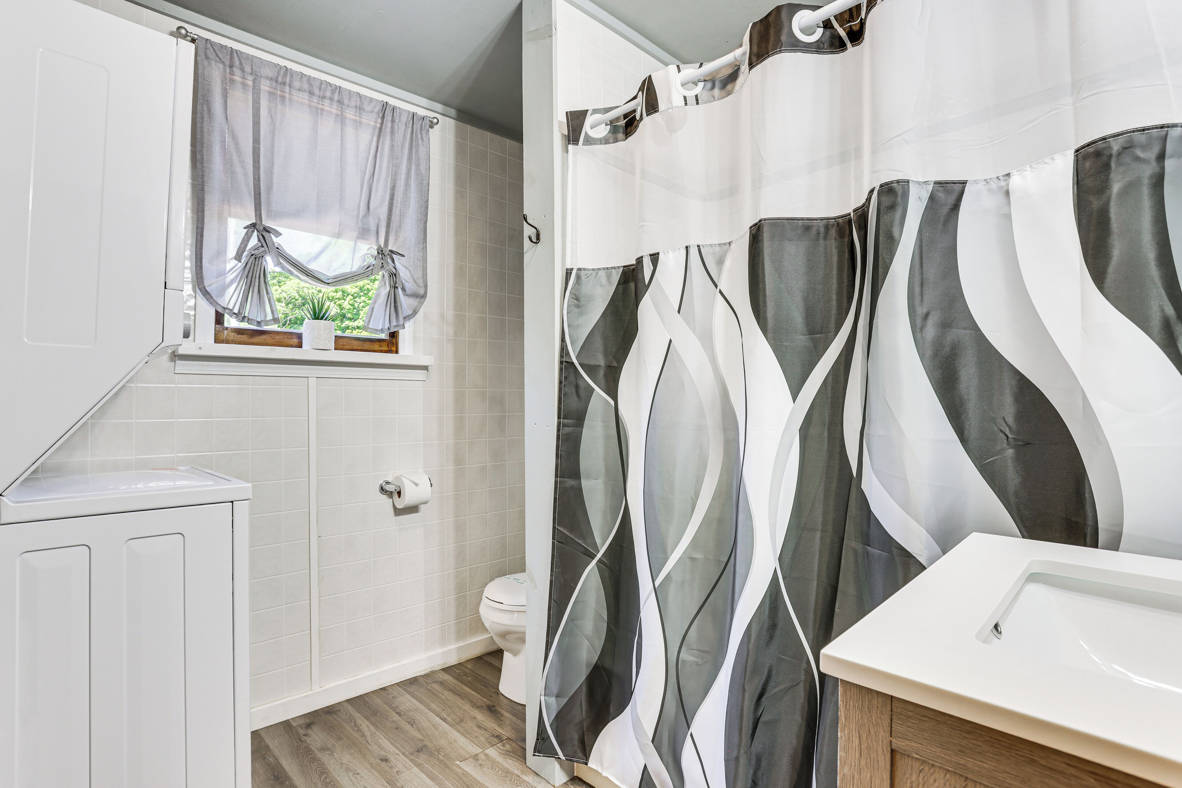A bathroom with a toilet, a sink, a window with curtains, and a shower curtain with a geometric pattern.