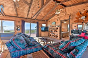 Interno di una baita in legno con un ampio divano, una televisione, una libreria e un balcone con vista.