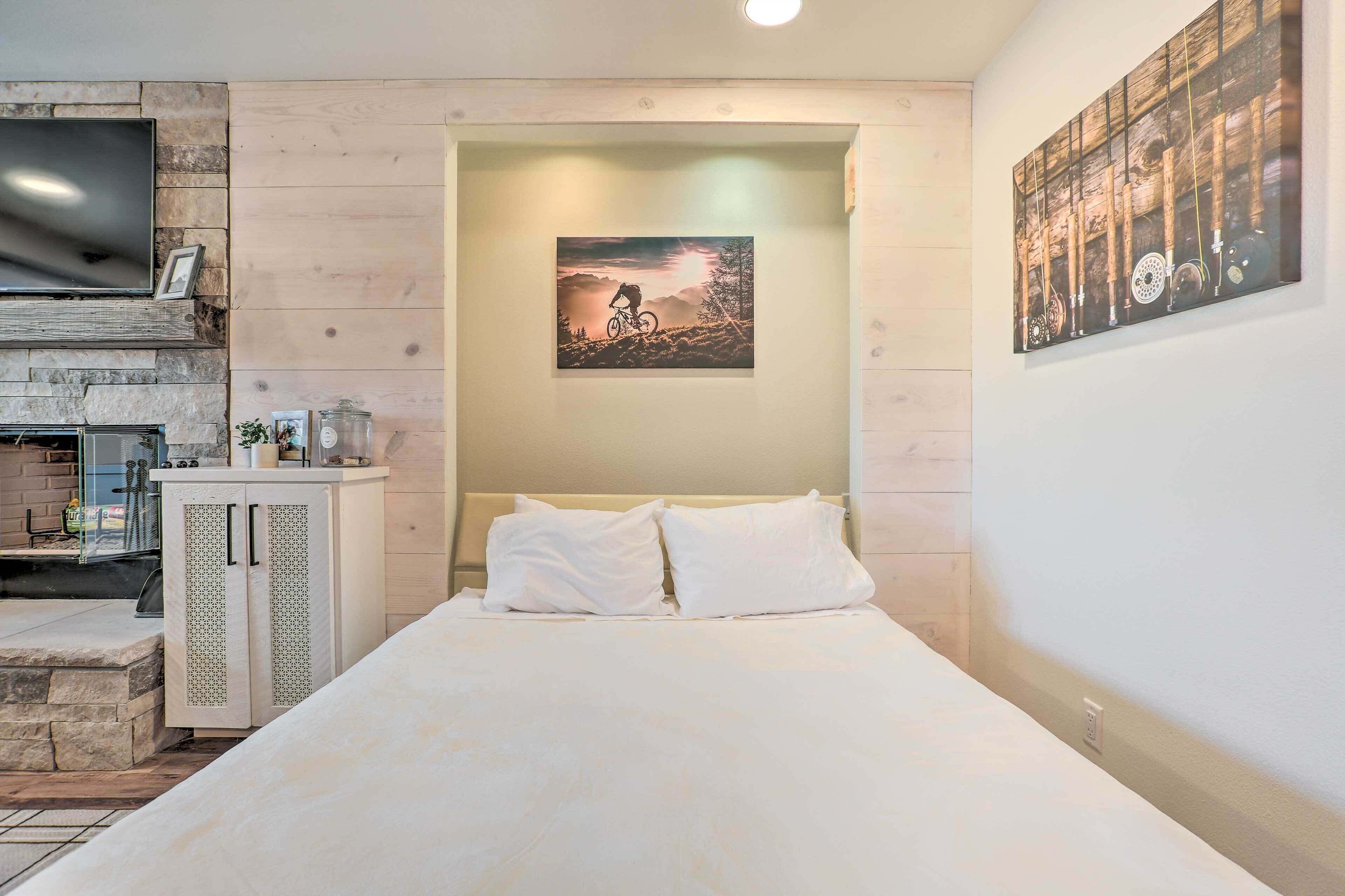 Un dormitorio con una cama, un televisor montado en una pared de piedra, un cuadro sobre la cama y un armario de madera con una mesita de noche al lado.