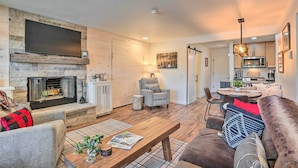 A living room with a fireplace, a flat-screen TV mounted on a stone wall, a dining area with a table and chairs, and a kitchen with modern appliances.