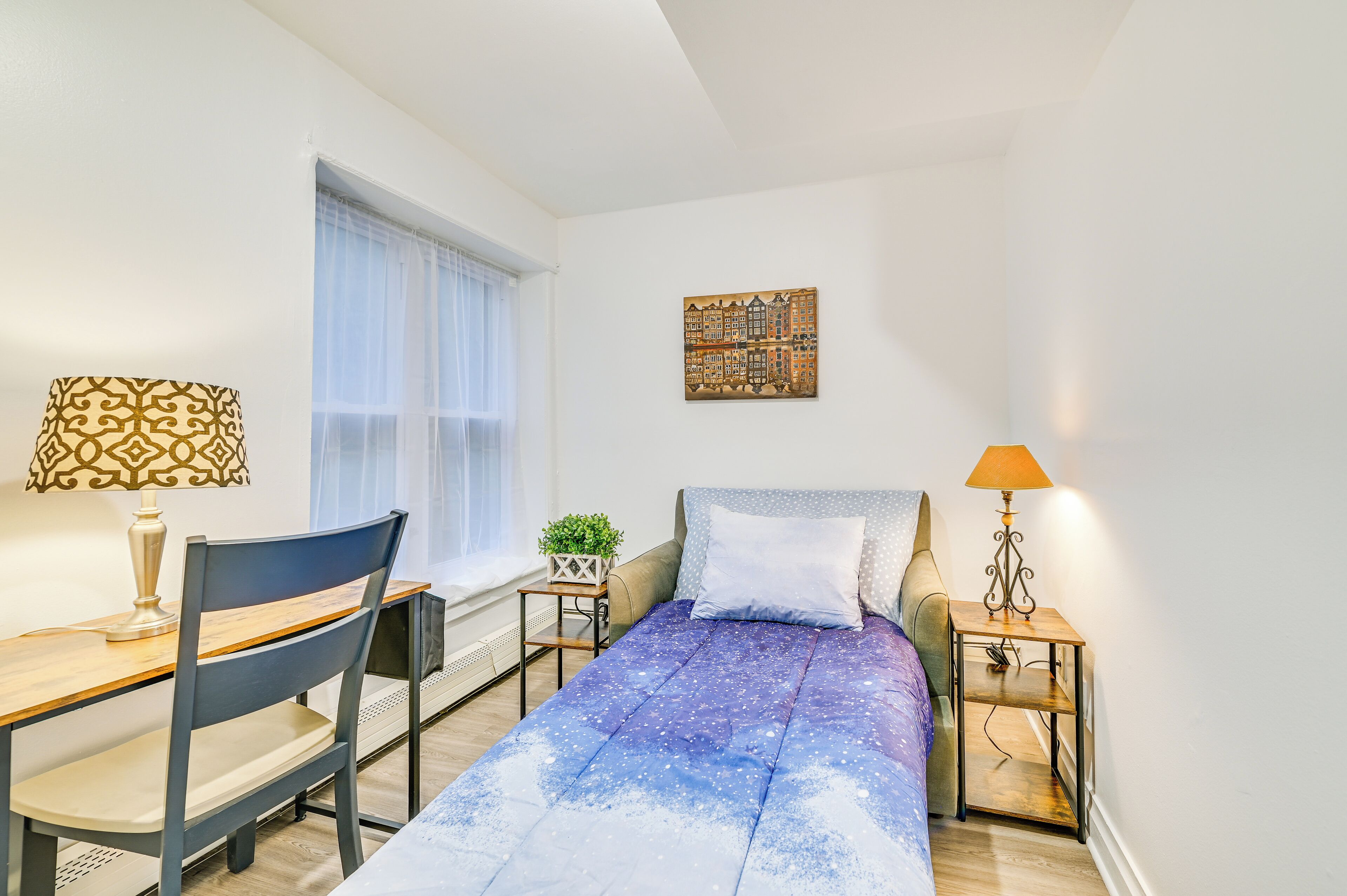 A small, well-lit bedroom with a bed, a desk, a chair, and a nightstand.