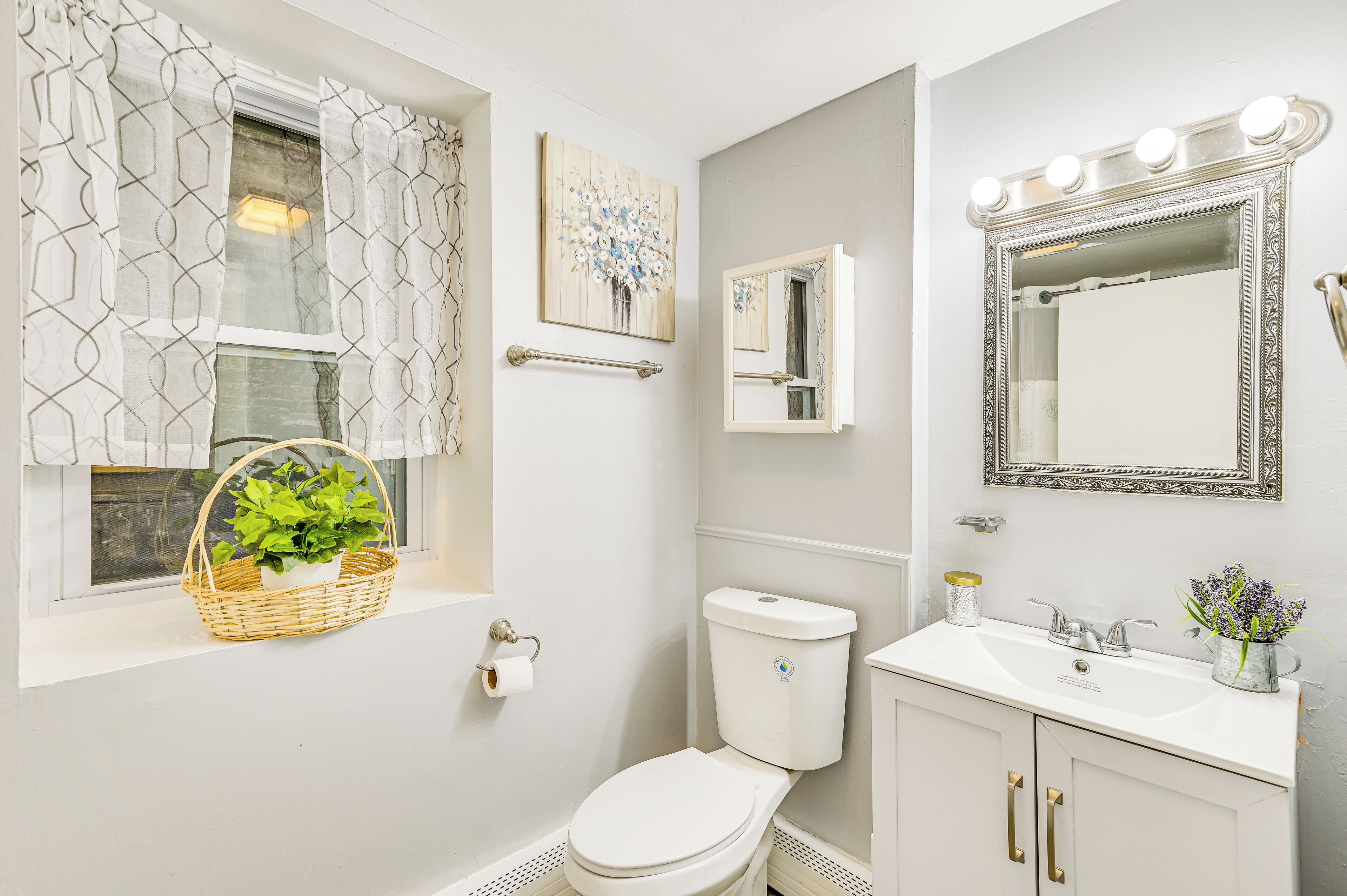 A bathroom with a toilet, sink, vanity, window with curtains, and a mirror with lights.