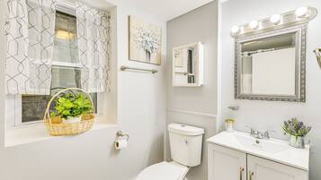 A bathroom with a toilet, sink, vanity, window with curtains, and a mirror with lights.