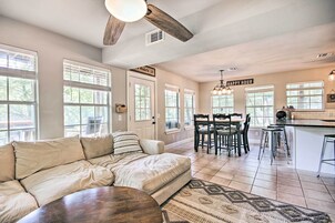 A modern living room with a beige sectional sofa, a wooden coffee table, and a patterned rug. There is a dining area with a wooden table and chairs, and a kitchen area with a bar and island.