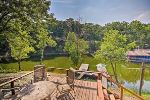 Une terrasse en bois, agrémentée de chaises et d’une table, offre une vue sur un lac paisible, le tout étant entouré d’une végétation luxuriante.