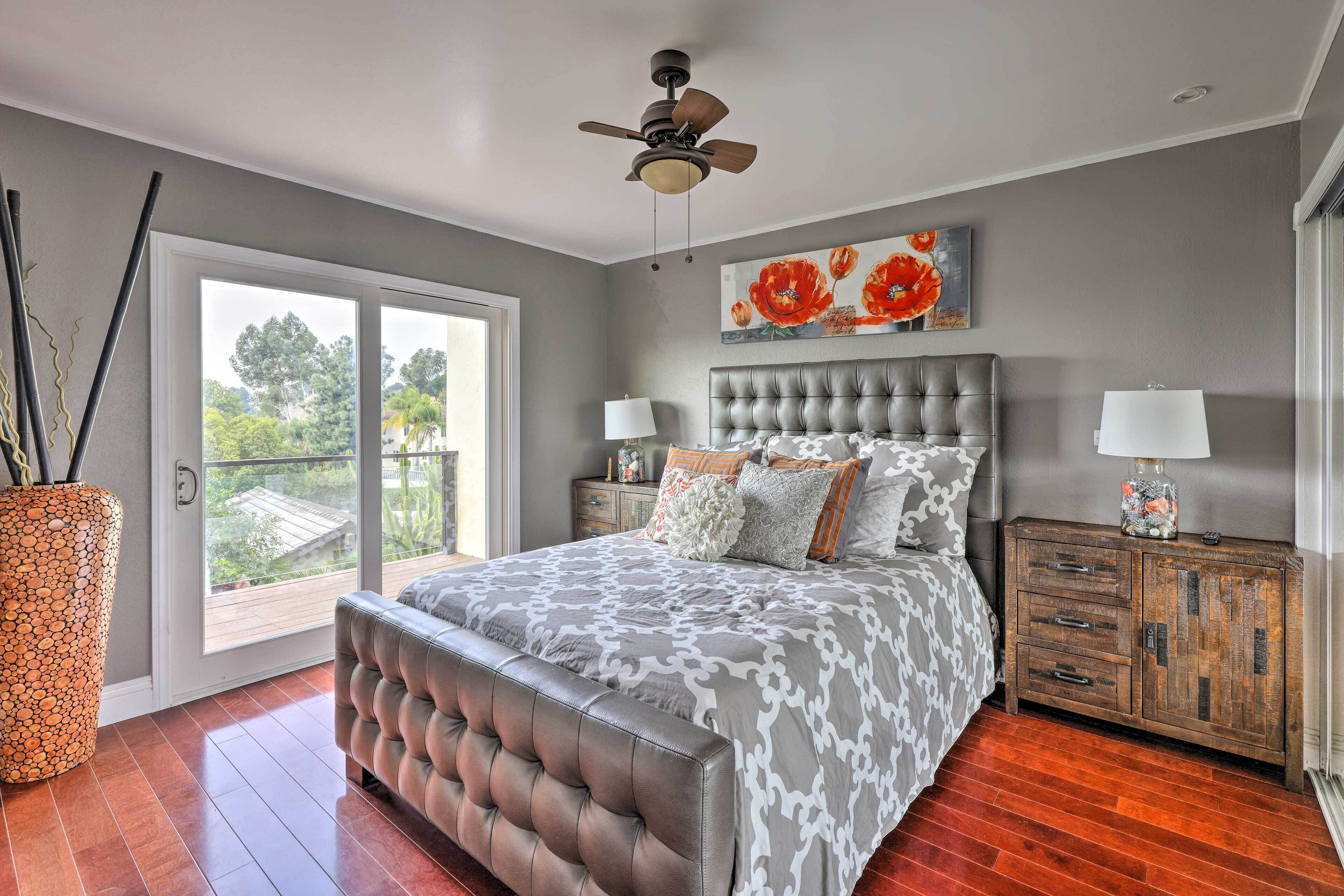 A bedroom with a large bed, a ceiling fan, a nightstand with a lamp, and a view of the outdoors.