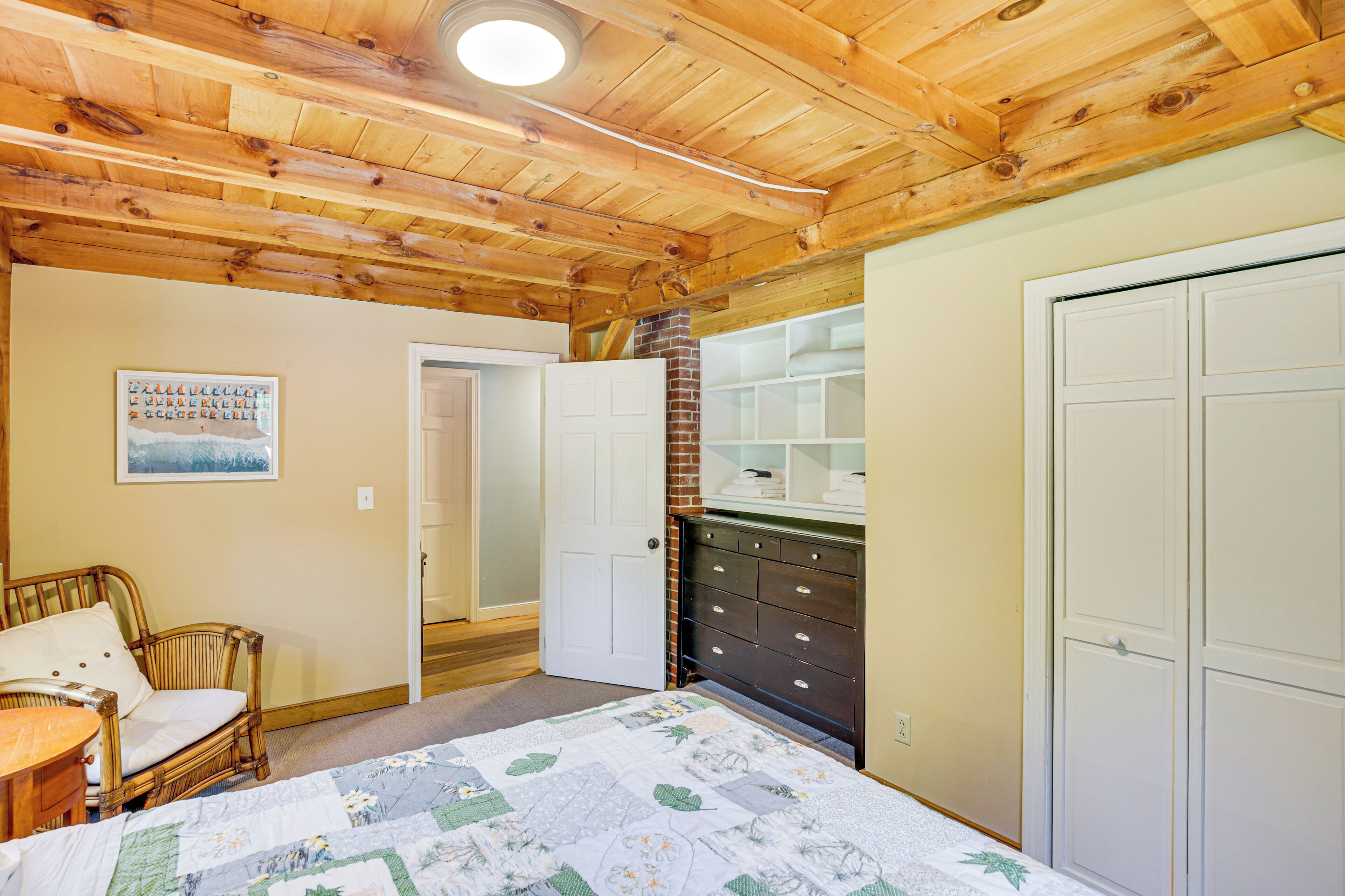 A bedroom with a wooden ceiling, a bed with a quilt, a dresser, a chair, and a door.