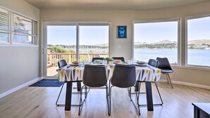 Un coin repas avec une table dressée pour quatre personnes, de grandes fenêtres offrant une vue sur un plan d’eau et des collines, et un balcon doté d’une balustrade en bois.