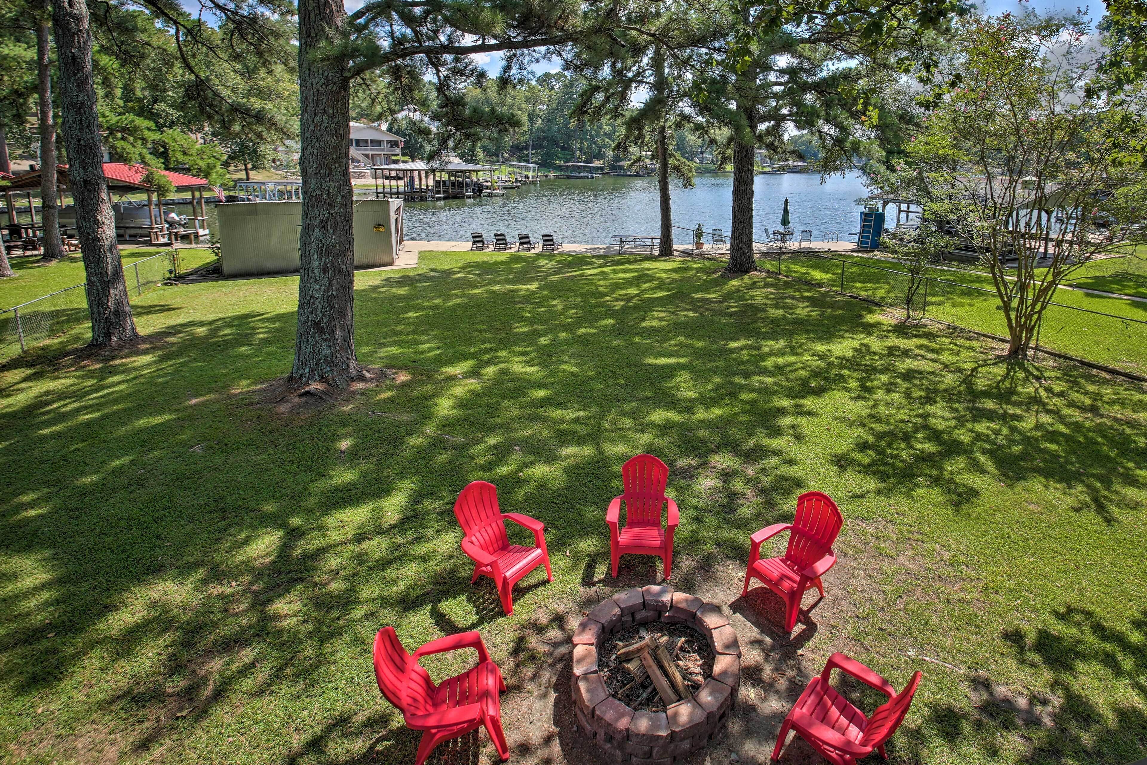 A grassy outdoor area with a fire pit and red chairs, overlooking a lake with docks and buildings in the background.