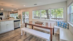 Une cuisine moderne avec des meubles blancs, une table à manger en bois et un banc. De grandes fenêtres laissent entrer la lumière naturelle et offrent une vue sur l’extérieur.