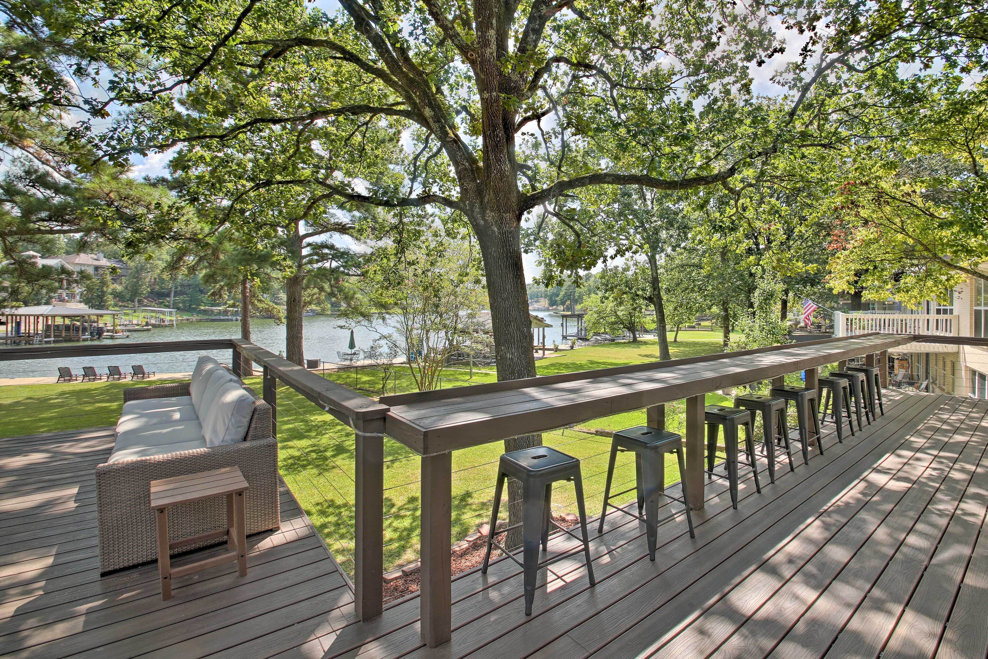 A wooden deck with a sofa and bar stools, overlooking a lake with a dock and chairs.