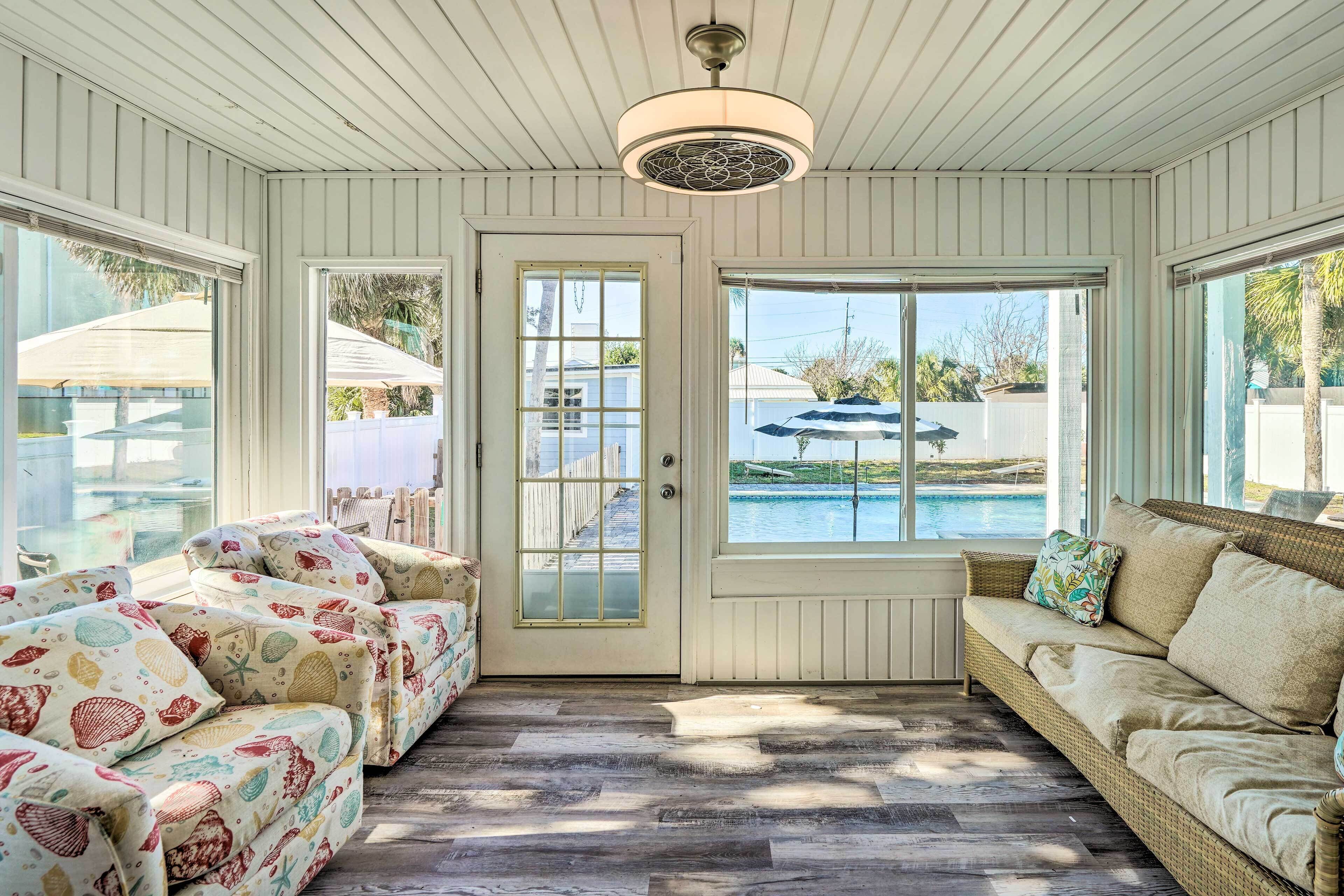 A sunlit room with a wicker sofa, a patterned armchair, and a view of a swimming pool.