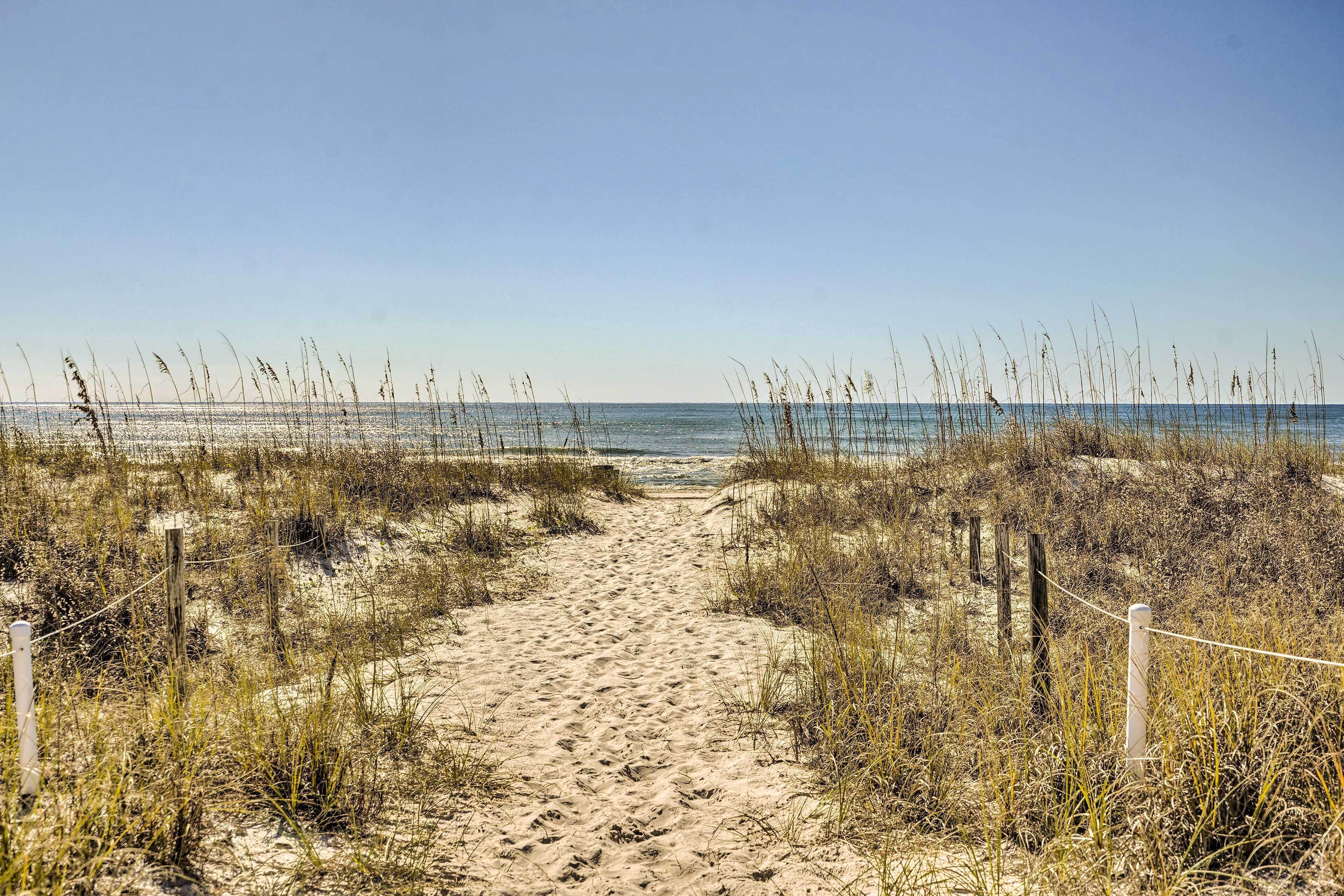 A sandy beach path leading to the ocean, surrounded by tall grasses and a wooden fence.