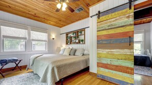 Ein Schlafzimmer mit einem Bett, einer Holzbalkendecke, einer Schiebetür im Bauernhausstil, einem Fenster und einem Regal mit Pflanzen.
