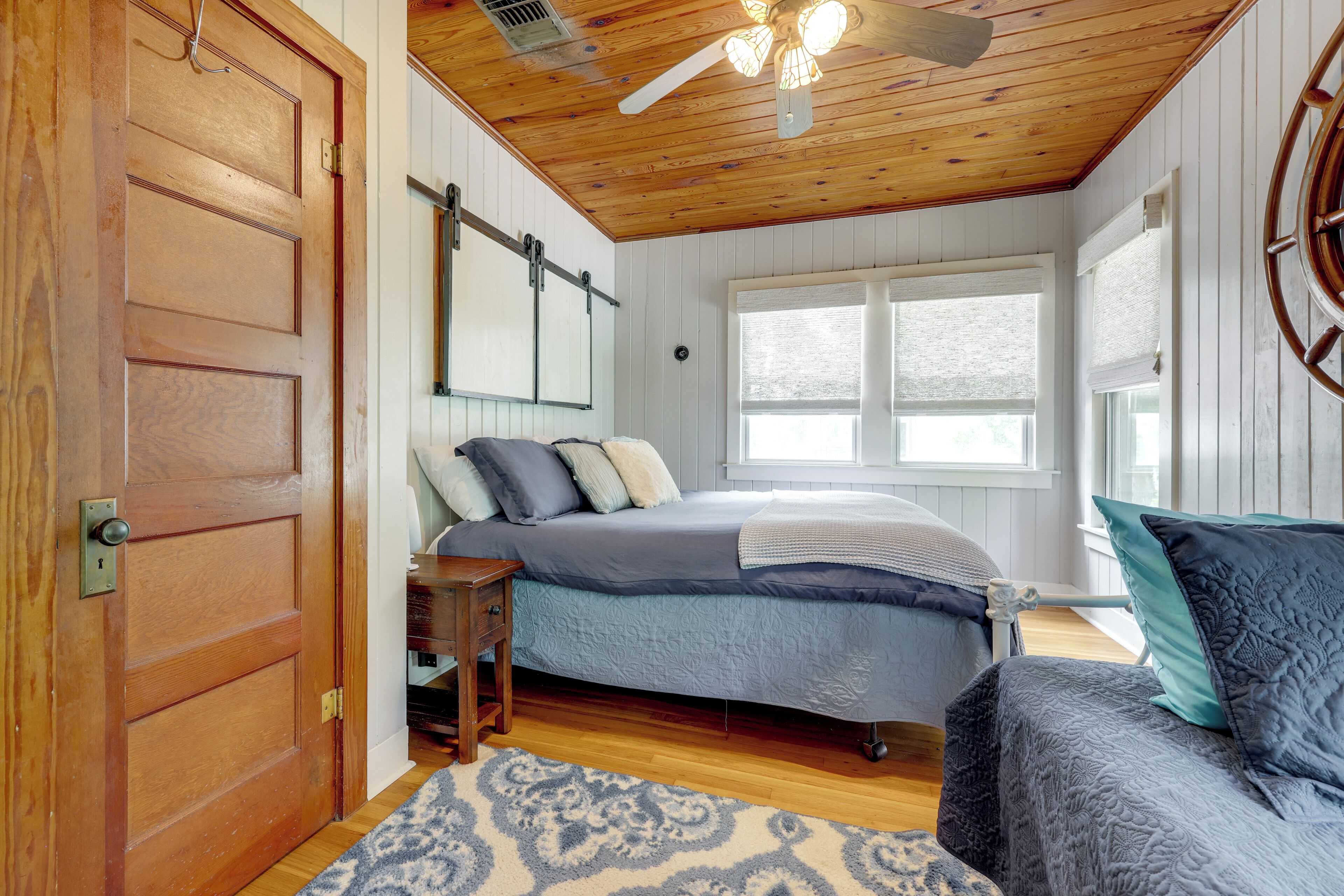 Une chambre avec un plafond en bois, un lit recouvert d’une courtepointe bleue, une table de chevet en bois et une fenêtre avec des stores.
