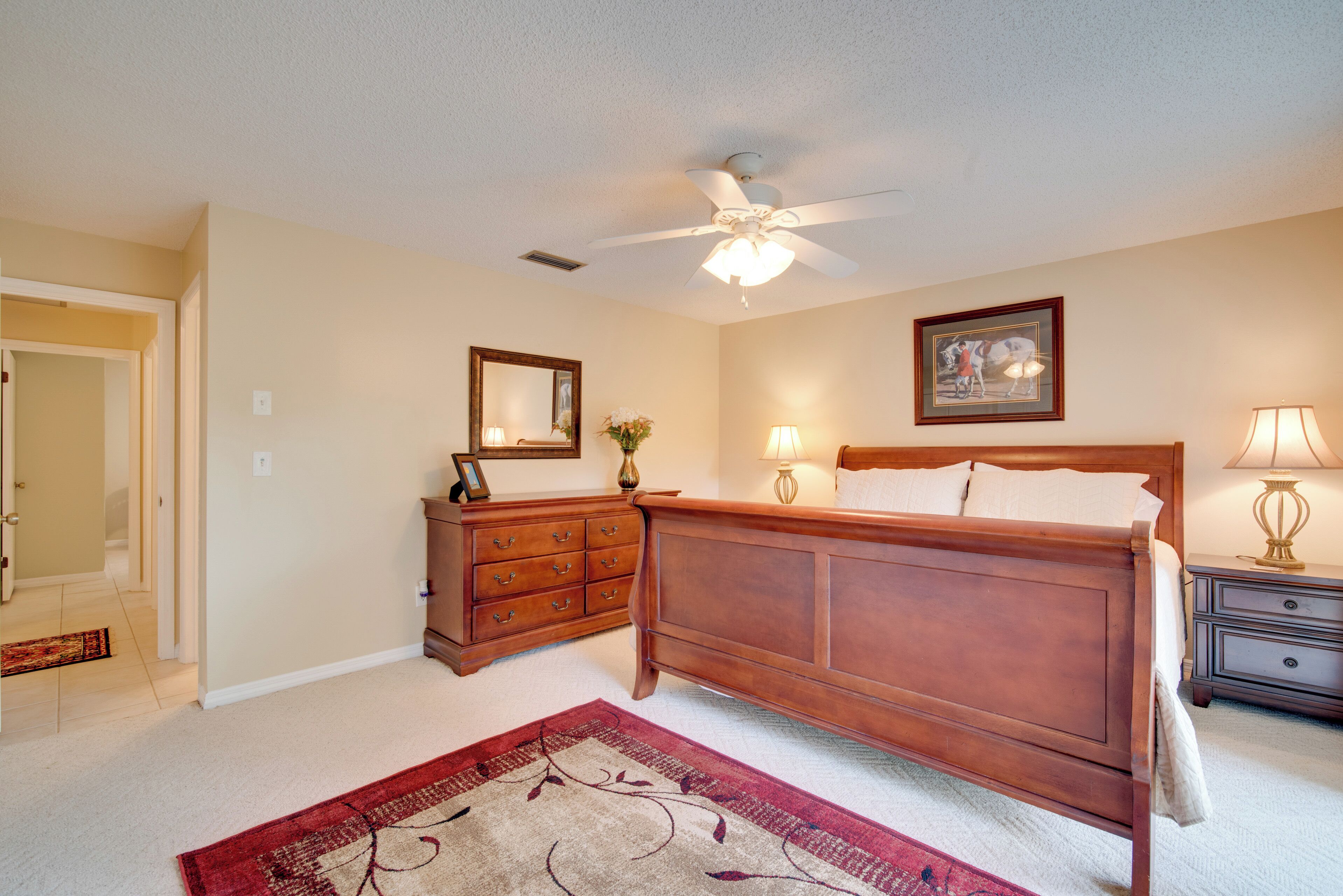 A bedroom with a wooden bed, a ceiling fan, a nightstand with a lamp, a dresser, and a framed picture on the wall.