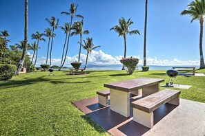 A grassy outdoor area with picnic tables and a barbecue grill, surrounded by palm trees and a view of the ocean. - Lush Molokai Island Oasis w/ Private Pool + Beach! (Kaunakakai)