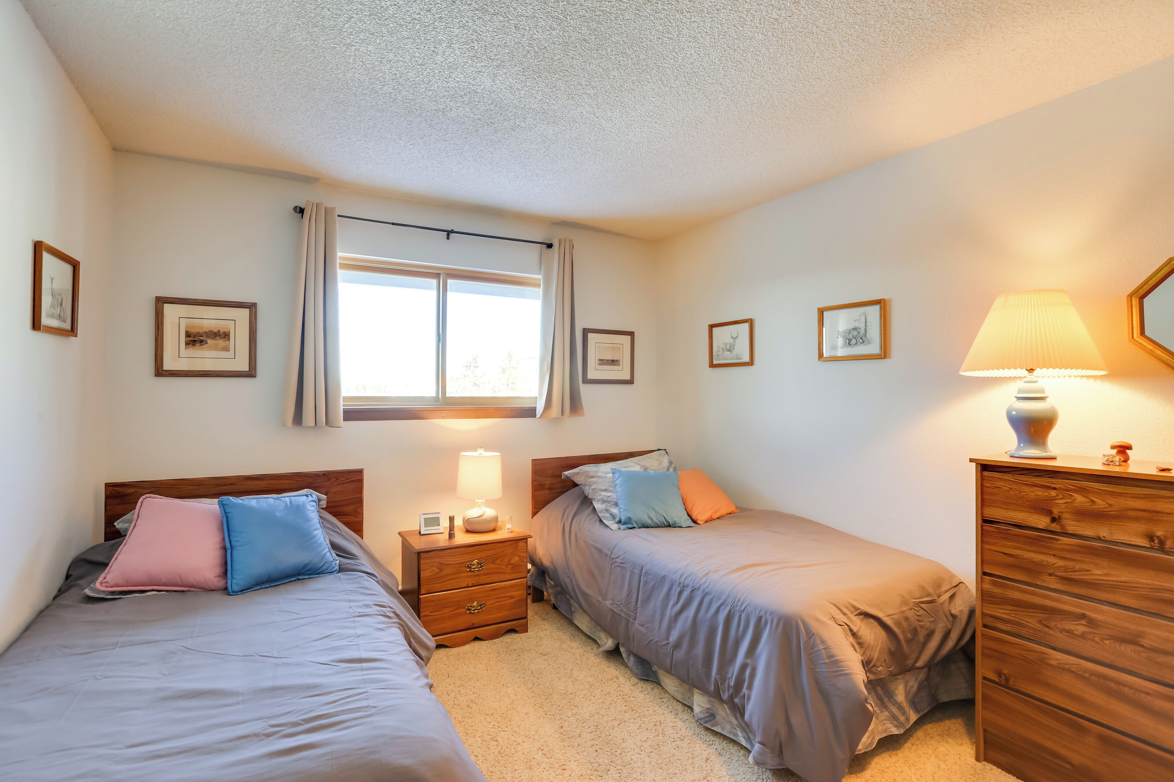 A bedroom with two beds, a nightstand with a lamp, a dresser, and framed pictures on the walls.