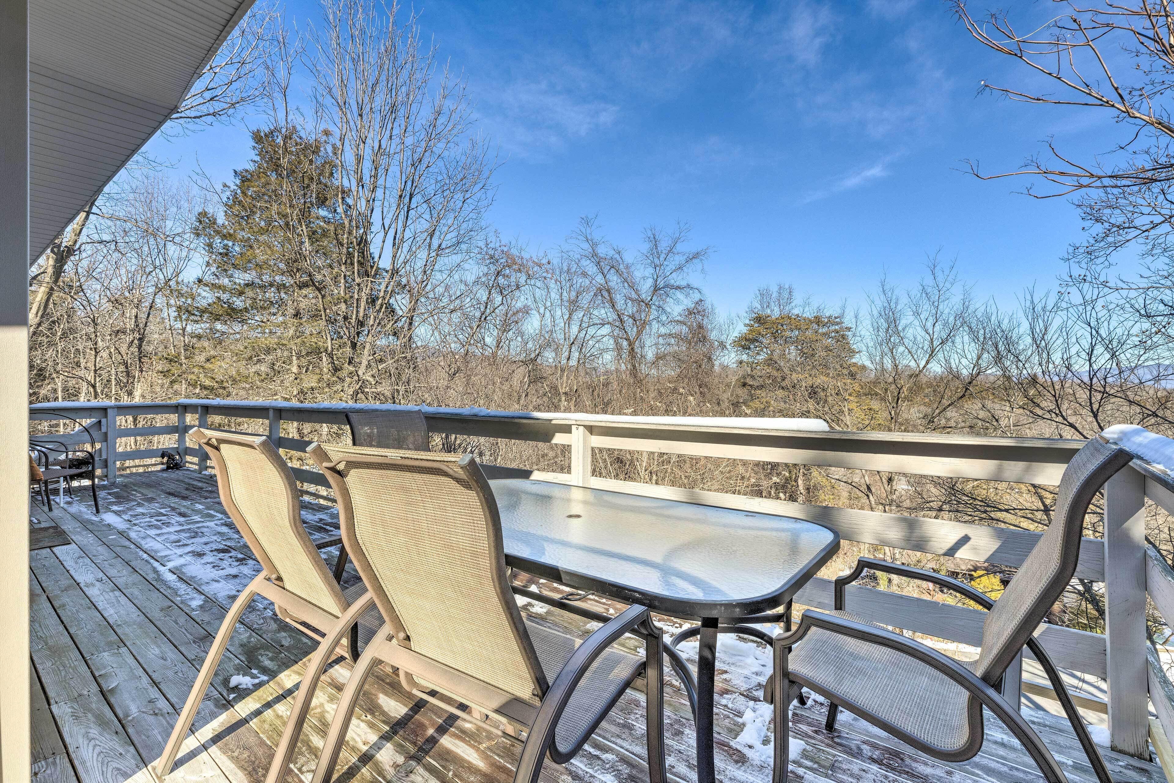 Una terraza de madera con sillas y una mesa de metal, con vista a un paisaje natural con árboles y cielo despejado.