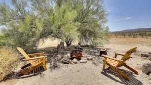 Two wooden Adirondack chairs, a metal fire pit, and a small table in a desert setting with sparse vegetation and distant mountains.