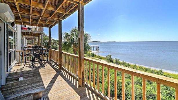 A wooden deck with a railing overlooking a body of water, a covered seating area with tables and chairs, and a view of the water. - Beachfront Cedar Key Condo w/ Pool, Spa & Views! (Cedar Key)