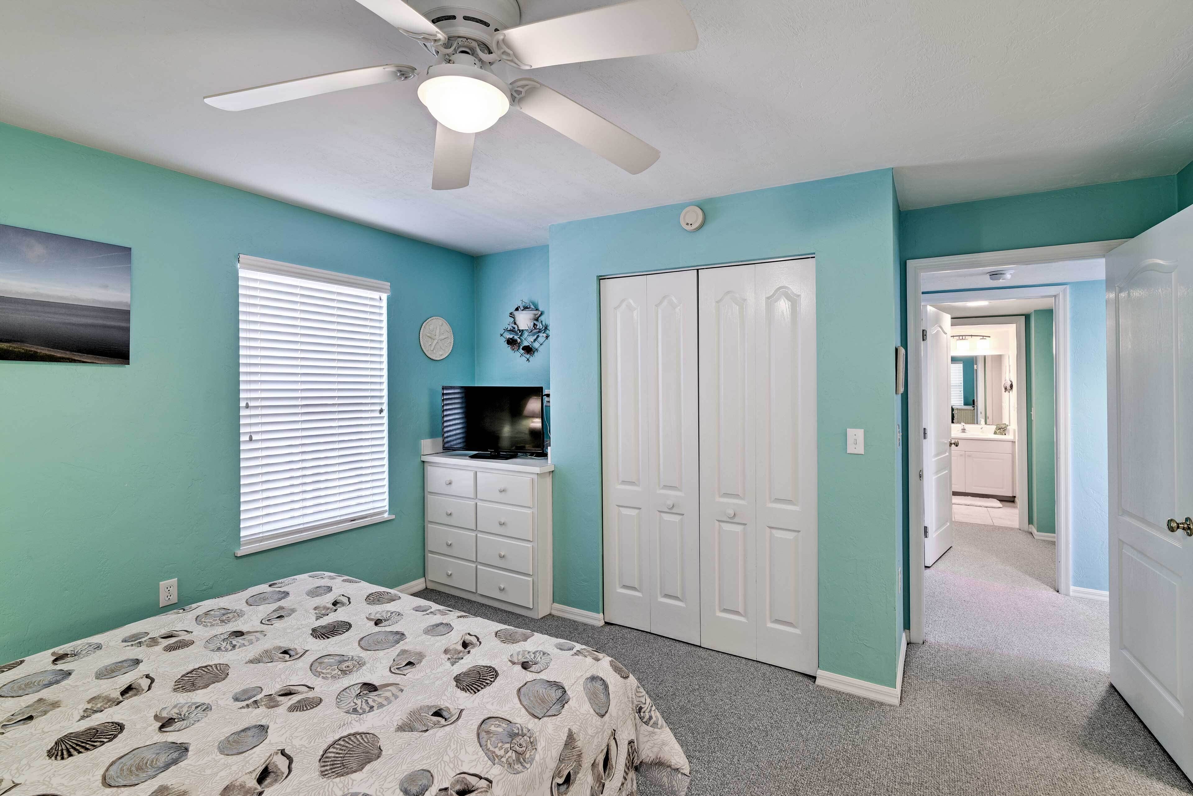 A bedroom with teal walls, a bed with a shell-patterned bedspread, a ceiling fan, a dresser, a TV, and a closet.