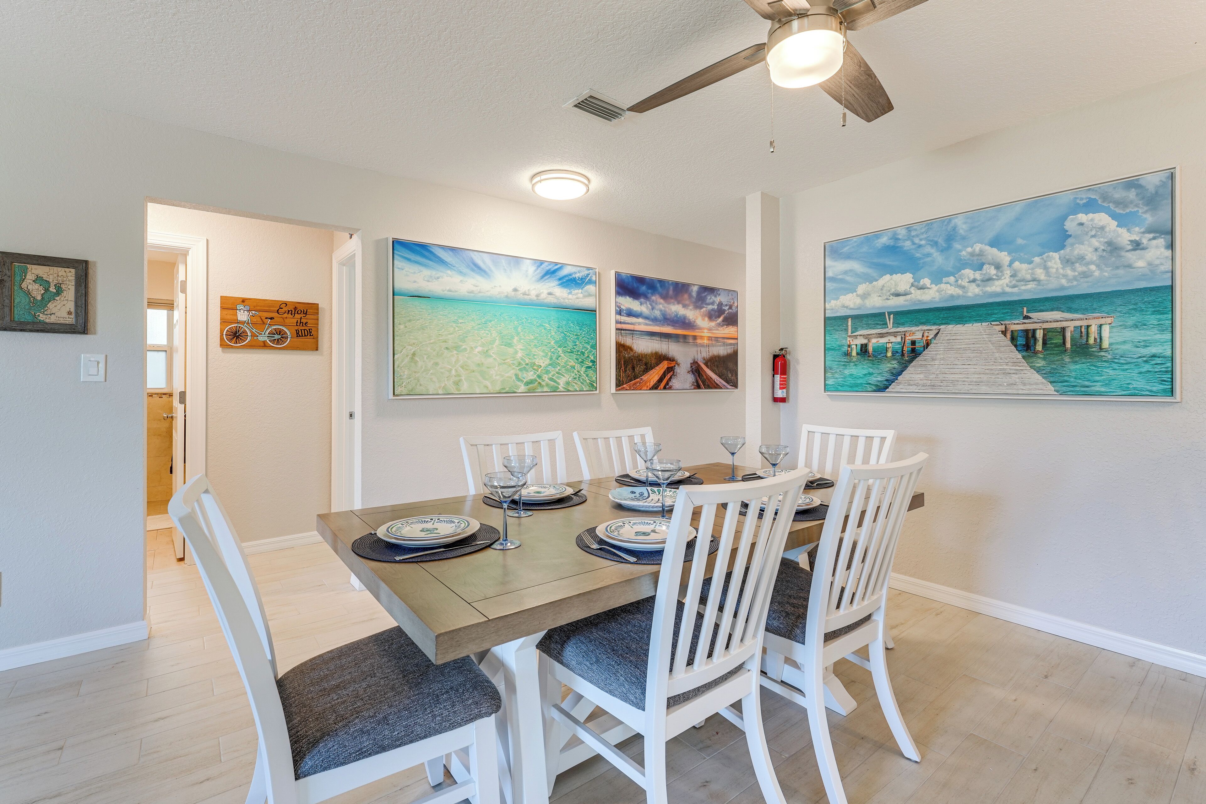 A dining area with a wooden table set for six, white chairs, and three framed paintings depicting tropical scenes.