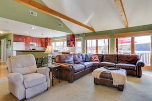 A spacious living room with a brown leather sectional sofa, a beige armchair, and a small ottoman. A wooden ceiling with exposed beams and large windows offering a view of the outdoors are visible.