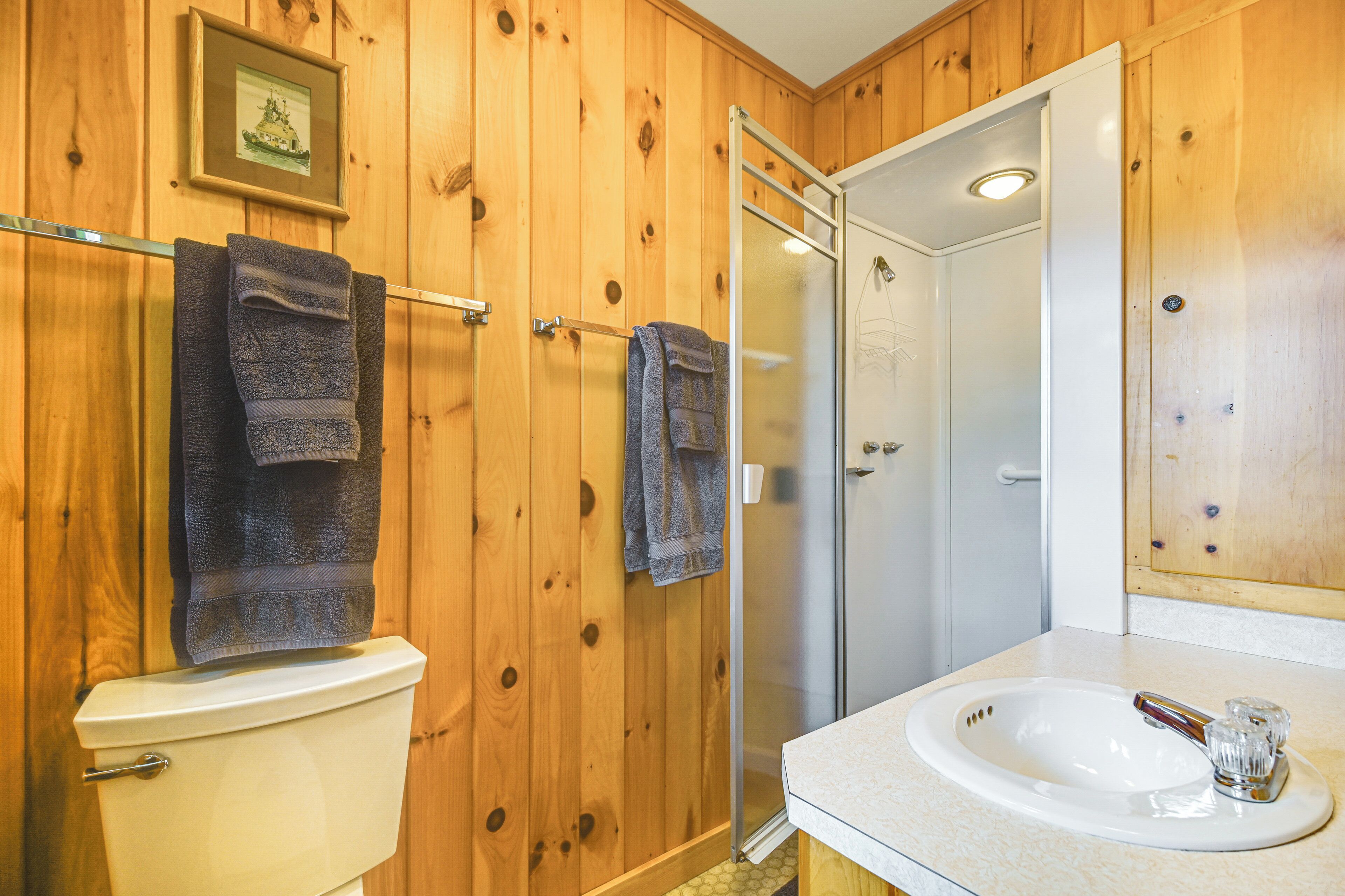Baño con paneles de madera, inodoro, dos toallas, cabina de ducha de vidrio y lavamanos con jarrón de vidrio.
