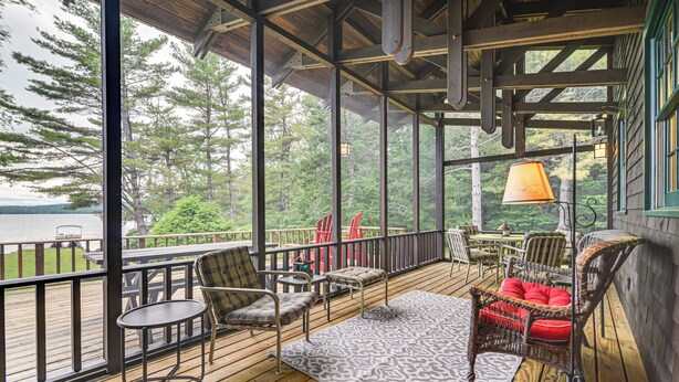 A wooden deck with outdoor furniture, a view of trees and water, and a covered area with a lamp.