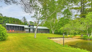 A modern house with a large deck, surrounded by lush greenery and a pond.